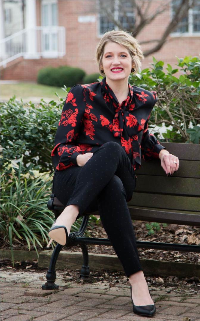Dr. Neiles sits on a bench wearing a black shirt with red flowers, black pants, and black high heels.